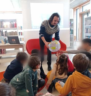 Atelier en médiathèque : Hugo Letellier tend un globe gonflable à des enfants assis en cercle autour de lui.