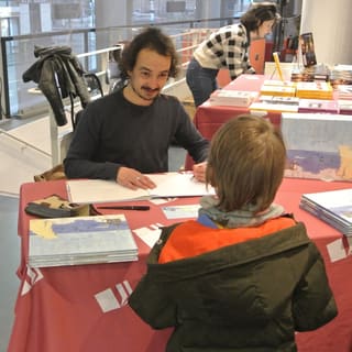 Séance de dédicace : Hugo Letellier dessine dans un album pour un jeune lecteur, à une table couverte de livres.