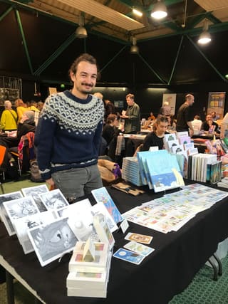 Hugo Letellier debout derrière son stand de salon du livre, garni d’albums, de cartes et d’illustrations.